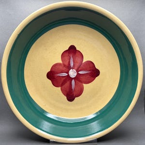 May include: A round ceramic serving bowl with a yellow interior and a dark green rim. The center features a red floral design with a white center. The bowl has a simple, rustic aesthetic.
