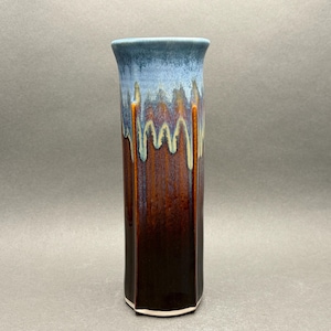 May include: A tall, cylindrical ceramic vase featuring a multi-toned glaze. The top section is a light blue that transitions into a deep, rich brown at the base, with vertical drip patterns creating a textured effect along the exterior surface.