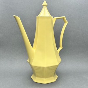 May include: A pale yellow coffee pot with a geometric design. The pot has a long spout, a handle, and a lid with a pointed top. The base of the pot has vertical ridges. The pot is made of ceramic or porcelain.