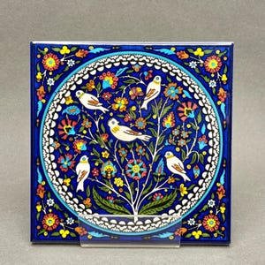 May include: A square ceramic tile featuring a central circular medallion with four white birds perched on a stylized tree. The design includes vibrant floral patterns in red, yellow, and light blue against a deep blue background with white borders.