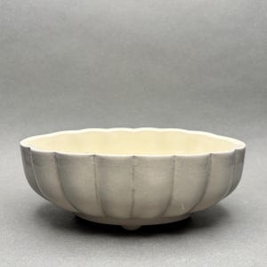 May include: A light grey ceramic bowl with a scalloped rim and a pale yellow interior. The bowl has vertical ridges on the exterior and three small feet. The bowl is a decorative piece.