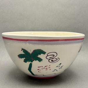 Può includere: Ciotola in ceramica color crema con bordo rosso e disegno di palma verde. La ciotola ha una striscia viola sotto il bordo e dettagli rossi alla base. La ciotola ha un tema tropicale.