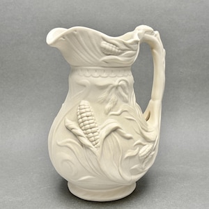 May include: Off-white ceramic pitcher with embossed corn and wheat stalk designs. The handle is shaped like a branch. The pitcher has a spout for pouring and a round base. A decorative piece for the kitchen or dining room.