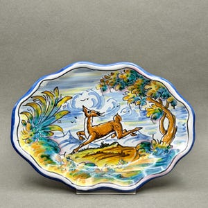 May include: An oval ceramic dish with a scalloped edge featuring a hand-painted scene of a brown deer leaping across a landscape with a tree, stylized foliage, and blue mountains under a cloudy sky, finished with a bold blue painted rim.