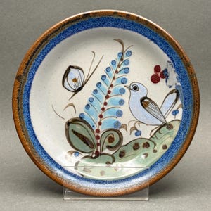 May include: A round ceramic plate with a cream-colored center and a wide, textured blue and brown rim. The center features a hand-painted design of a light blue bird, a butterfly, a tall blue floral stalk, and green leaves with brown spots.