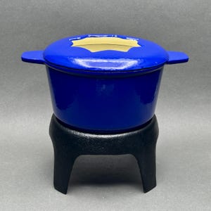 May include: A cobalt blue cast iron fondue pot with a matching lid and a black stand. The pot has two side handles and a light yellow interior. The stand has a three-legged design.