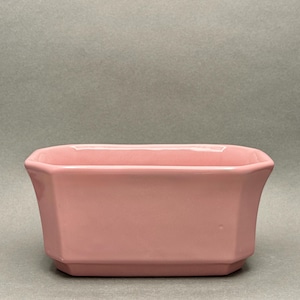 May include: A rectangular, light pink ceramic planter with a geometric design. The planter has a slightly flared shape and a smooth, glossy finish. The planter is suitable for small plants or succulents.