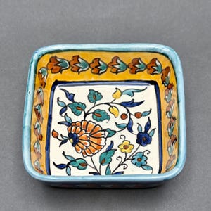 May include: Square ceramic dish with a floral design. The dish features a white center with an orange and blue floral pattern. The border is yellow with orange and green floral accents, and a light blue rim.