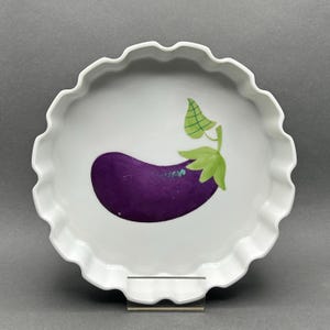May include: White ceramic serving dish with a scalloped edge. The dish features a hand-painted purple eggplant with green leaves. The design is centered on the white surface. The dish is on a clear stand.