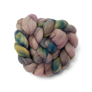 May include: A close-up of a braided yarn skein in shades of pink, grey, blue, and yellow. The yarn is thick and textured, with a soft, fluffy appearance. The skein is arranged in a circular shape against a white background.