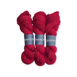 May include: Three skeins of vibrant red hand-dyed yarn are shown against a white backdrop. Each skein is wrapped with a blue label that reads "Cheshire Yarn Hand Dyed". The yarn is thick and appears soft.