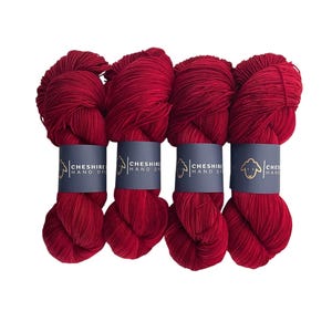 May include: Four skeins of deep red yarn, each with a navy blue label displaying a sheep logo and the text "Cheshire Hand Dyed." The yarn is tightly wound, presenting a rich, solid colour, ideal for knitting or crochet projects.