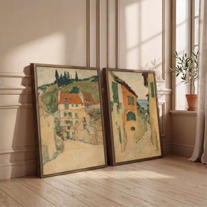 May include: Two framed rectangular wall art prints featuring impressionist-style paintings of European village landscapes. The artworks display buildings with orange roofs, green shutters, and stone walls, set against rolling hills and trees.