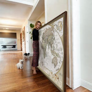 A woman stands in a room, leaning against a large world map, with two small dogs sitting on the floor in front of her.
