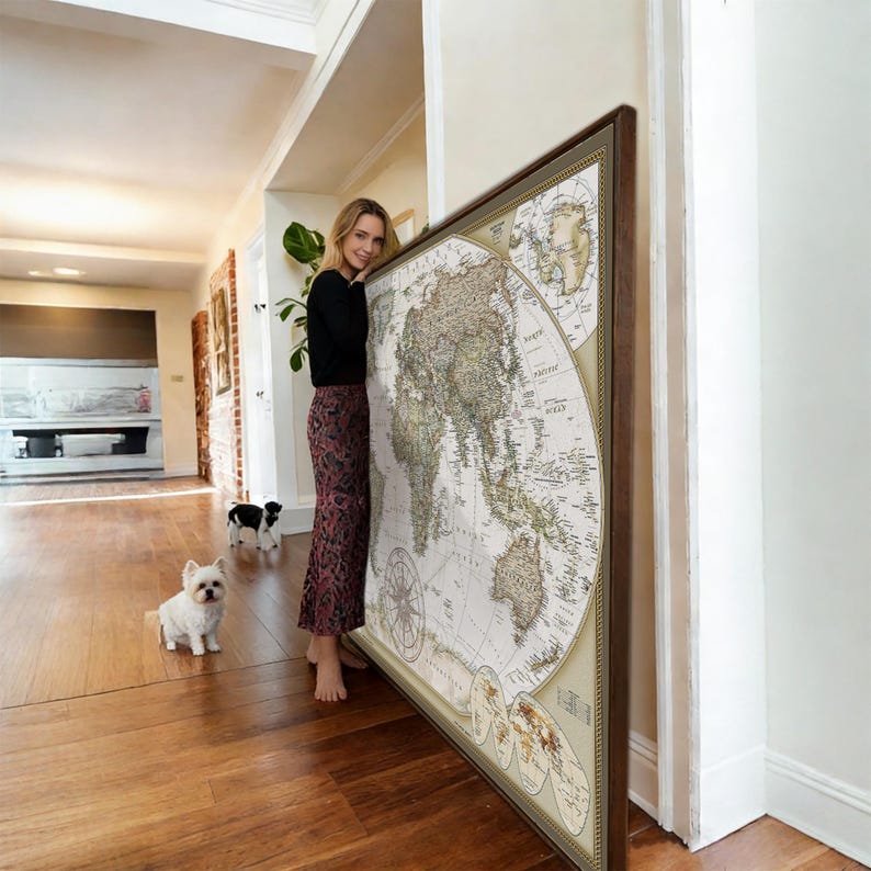 A woman stands in a room, leaning against a large world map, with two small dogs sitting on the floor in front of her.