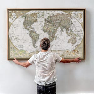 May include: A framed vintage world map with a detailed cartographic design in muted tones of beige, green, and brown. The map is held by a person, showcasing its large rectangular shape and wooden frame.