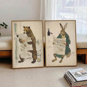 May include: Two framed rectangular wall art prints featuring anthropomorphic animals. The left print shows a brown bear reading a newspaper in a bathroom. The right print depicts a brown rabbit wearing a green coat and reading a newspaper.