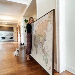 May include: A large, rectangular wall map of the world featuring a cream-coloured background with continents depicted in muted shades of orange, green, and blue, all enclosed within a dark brown wooden frame and displayed in a vertical orientation.