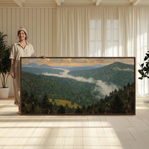 May include: A large rectangular landscape painting in a dark wood frame depicting a dense forest valley at sunrise. The scene features rolling hills covered in deep green trees, with wispy white mist settling in the lowlands and a soft orange sky.
