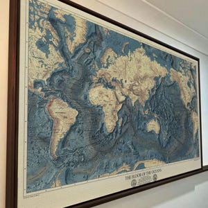 Puede incluir: Un gran mapa mural rectangular titulado THE FLOOR OF THE OCEANS, enmarcado en madera oscura. El mapa muestra la batimetría global con fosas oceánicas de color azul profundo y masas terrestres beige, con topografía submarina detallada.