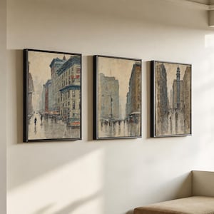 May include: A set of three framed vertical wall art prints depicting urban city street scenes in a muted color palette. Each rectangular piece features tall buildings, pedestrians, and rainy weather conditions rendered in a painterly style.