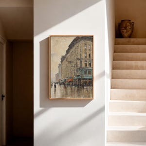 May include: A framed painting depicts a European street scene with a large building and shops. The artwork features muted tones of beige, brown, and gray, with a wet, reflective street. People with umbrellas are walking on the street.