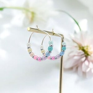 Könnte beinhalten: Ein Paar Creolen mit bunten Perlen. Die Ohrringe befinden sich auf einem Holzständer und werden vor einem weißen Hintergrund präsentiert.