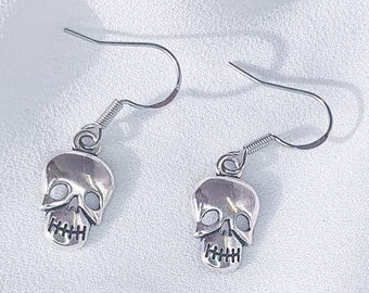Puede incluir: Un par de pendientes de calavera plateados con un diseño sencillo. Los pendientes están sobre un fondo blanco.