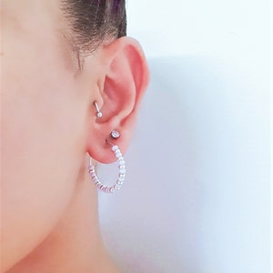 May include: A close-up of a person's ear with a silver hoop earring made of small white and purple beads. The earring is in the lower lobe of the ear. There is a small silver stud earring in the upper lobe of the ear.