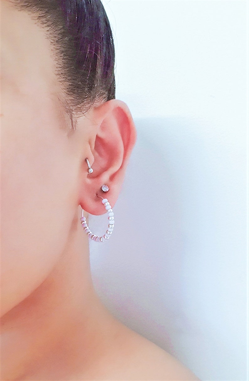 May include: A close-up of a person's ear with a silver hoop earring made of small white and purple beads. The earring is in the lower lobe of the ear. There is a small silver stud earring in the upper lobe of the ear.