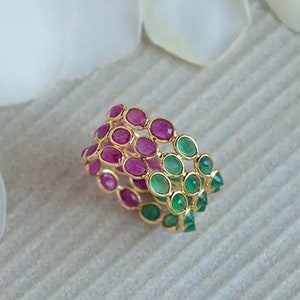 Pode incluir: Um anel de ouro com um design graduado de pedras preciosas rosa e verdes. O anel está sobre um fundo branco.