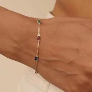 May include: A gold bracelet with a delicate chain and rectangular gemstones in green, red, and blue. A matching gold ring with colorful gemstones is also visible. The jewelry is worn on a person's wrist and finger.