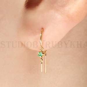 May include: A close-up of a gold earring with a small green gemstone and two gold bars hanging below. A gold ear cuff is also visible. The earring is attached to the ear. The text "GEMSTUDIOBYRUBYKHAN" is visible.
