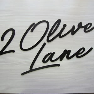 May include: Black cursive script house number sign with the number "2" and the street name "Oliver Lane".