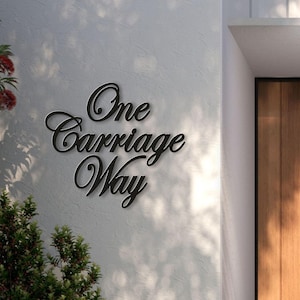 May include: Black metal sign with the words "One Carriage Way" in a cursive font, mounted on a textured white wall. The sign is in a shadow, with greenery in the foreground.