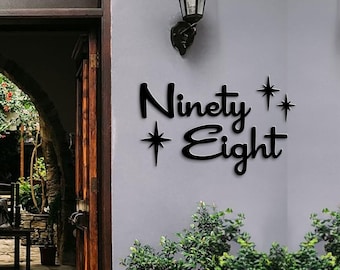 May include: Black metal sign with the text "Ninety Eight" and two stylized stars on a white wall.