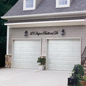 May include: A black metal address sign mounted on the exterior wall above two white garage doors. The sign features the text 119 Major Buttons Dr in a cursive script font. Two black outdoor wall lanterns are positioned on either side of the sign.