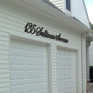 May include: A black metal house sign mounted above a white garage door, featuring the text 135 Sullivan Avenue in a cursive script font. The sign is attached to the white horizontal siding of a building exterior, displaying the address clearly.