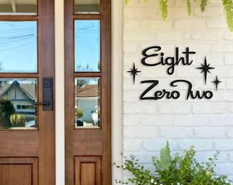 May include: Black metal house number sign that reads "Eight Zero Two" with starburst accents. The sign is mounted on a white brick wall near a wooden door with glass panels.