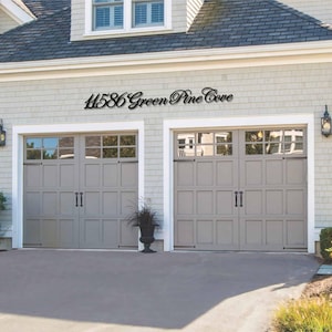 May include: A black metal wall sign featuring the address 11586 Green Pine Cove in a cursive script font. The sign is mounted on the light gray shingled exterior wall of a house, positioned directly above two gray garage doors with window panels.