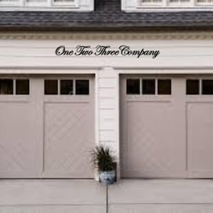 Peut inclure: Une maison blanche avec deux portes de garage. Les portes de garage sont beiges avec un motif en losange et ont des fenêtres. Le texte "One Two Three Company" est au-dessus des portes de garage.