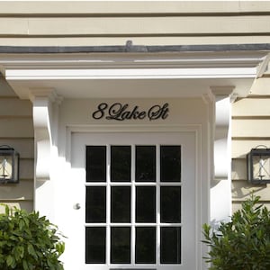 May include: A white front door with a grid of rectangular glass panes. Above the door, the black script reads "8 Lake St". The door is framed by white trim and flanked by two black lanterns and green bushes.