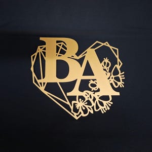 May include: A gold-coloured laser-cut sign featuring the letters BA in a bold serif font. The letters are framed by a geometric heart shape outline and a decorative floral arrangement on the right side, creating a flat, cutout design.