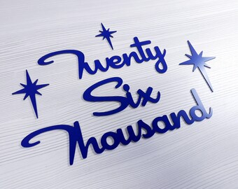 May include: Blue decorative cutouts spelling out "Twenty Six Thousand" with star accents. The lettering is in a cursive font, and the stars have a pointed design. The cutouts are placed on a white, wood-grain surface.