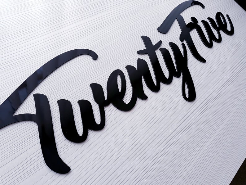 May include: Black acrylic cutout of the word "Twenty Five" in a cursive font.