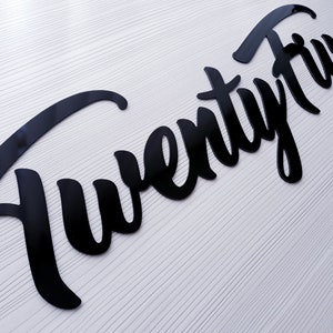 May include: Black cursive script lettering that reads "Twenty Five" on a white background.