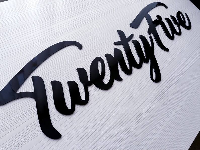 May include: Black cursive script lettering that reads "Twenty Five" on a white background.