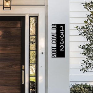 May include: A vertical rectangular black metal house address sign featuring the text PORT COVE DR on the left side and the numbers 1332 on the right side, mounted on a white wall next to a dark wood front door with a glass side panel.