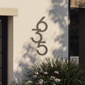 May include: Bronze-colored house numbers 635 mounted on a light-colored wall. The numbers are in a modern, sans-serif font and are vertically aligned. The numbers are displayed outside a building, with greenery visible at the bottom.