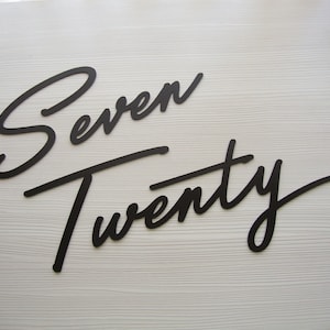 May include: Black script font cutouts of the words "Seven Twenty" on a white wood background.